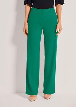 Vrouw draagt groene broek met hoge taille en wijde pijpen met riemlussen, gecombineerd met nude puntige hakken en een zwarte top, staand tegen een effen achtergrond.