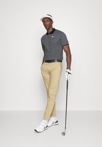 Randig svart och vit polo skjorta, beige byxor, vita sportskor, golfhandske, håller i en golfklubba, bär en vit visor.