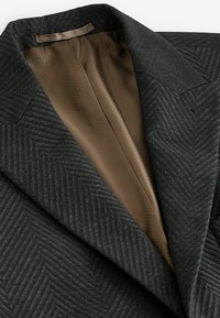 Czarna marynarka w jodełkę z teksturowaną powierzchnią, ostrymi klapami i brązową satynową podszewką. Strukturalny krój z detalicznym przeszyciem.