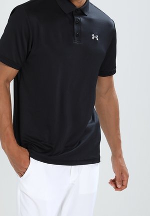 Man draagt een zwart poloshirt met korte mouwen met een klein logo op de borst en een witte broek, staat met één hand in de zak.