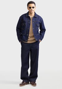 Jeansjacke mit traditionellen Taschen und braunen Nähten, kombiniert mit einem beigem Kabelstrickpullover und lockeren Jeanshosen. Braune Schuhe.