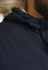 Marineblauwe parka met nep bont aan de kap, voorzien van trekkoordverstellingen en een gestructureerde stof. Close-up van het kraaggebied.