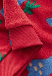 Roter Fleece-Stoff mit einem blauen Hintergrund und grünen sowie gelben Mustern. Der Ärmelabschluss ist gerippt und bietet einen strukturierten Kontrast.