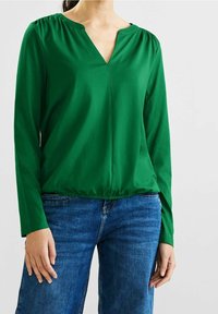 Grön långärmad topp med V-ringning och samlad axeldetalj, har en lätt elastisk avslutning vid nederkanten, i kombination med blå jeans.