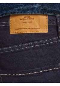 Mörkblå denimjeans med en ljusläderlapp med präglad text. Guldstickningar accentuerar jeansens bakfickor och sömmar.