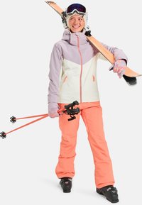 Ski outfit featuring a light purple and cream jacket with orange accents, orange pants, pink gloves, and skis with black bindings.