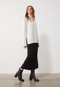 Un pull à col roulé blanc côtelé associé à une jupe midi en tricot noir et des bottines à talons noires, sur un fond clair.