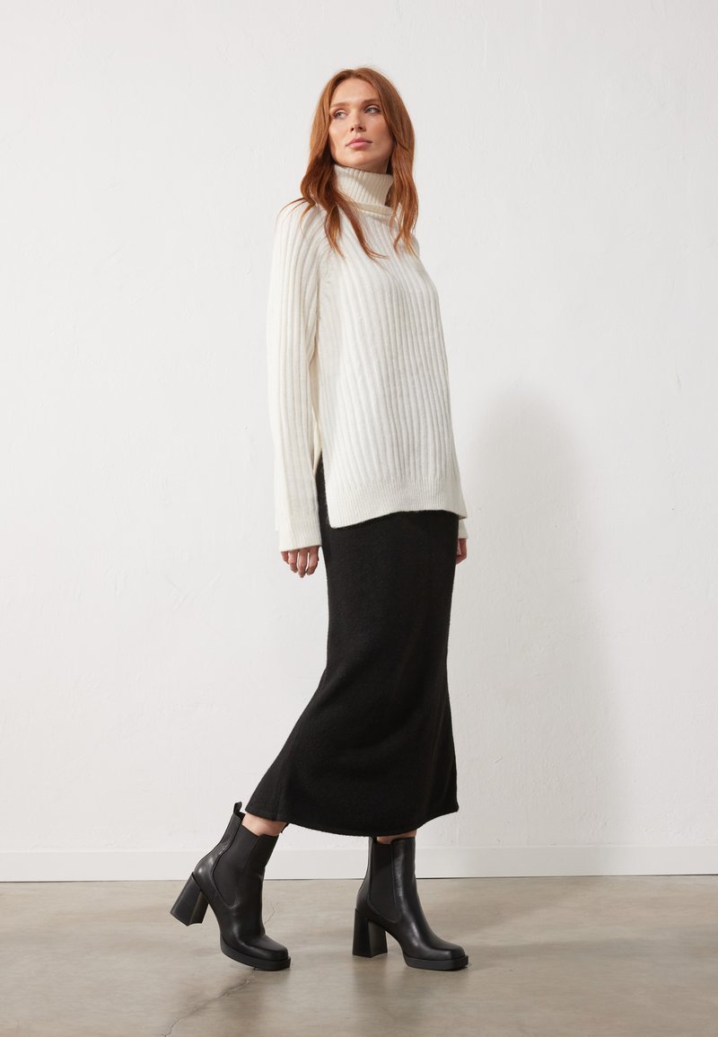 Un pull à col roulé blanc côtelé associé à une jupe midi en tricot noir et des bottines à talons noires, sur un fond clair.