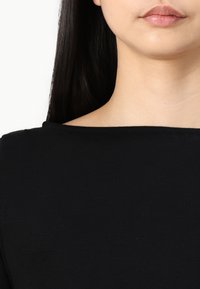 Fekete felső, kerek nyakkivágással és sima textúrával, egyszerű, testhezálló kialakítással, amely hangsúlyozza a kulcscsontot és a vállakat.