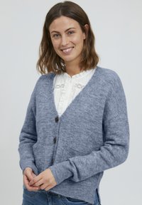 Gai pelēks kardigāns, izgatavots no mīksta vilnas maisījuma, ar V-veida kakla izgriezumu, divām priekšējām kabatām un trim brūnām pogām.