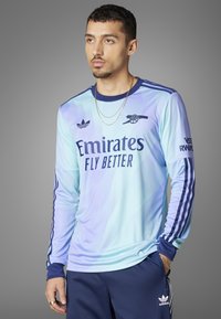 Lyseblå langærmet trøje har et gradientdesign, marineblå detaljer og "Emirates Fly Better" logo på forsiden. Adidas-striber på ærmerne.