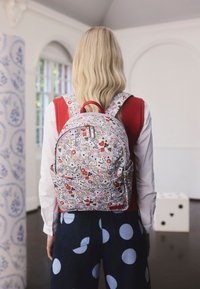 Szőke nő mintás hátizsákkal, amely játékkártyákat és kockákat ábrázol, piros mellényben, fehér ingben és kék pöttyös nadrágban.