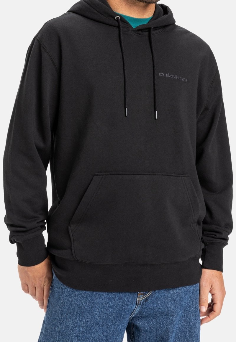 Uomo che indossa un hoodie nero Quiksilver con tasca frontale e jeans blu, inquadrato all'altezza del mento e dei fianchi, in piedi su uno sfondo bianco.