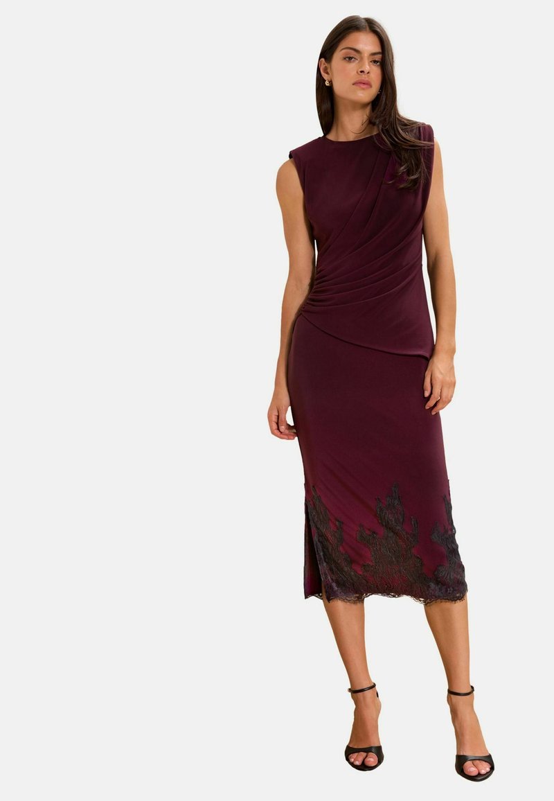 Woman wearing a sleeveless burgundy midi dress with side pleats and black lace detailing at the hem, paired with black ankle-strap heels.