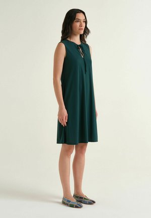 Donna in piedi che indossa un abito senza maniche verde scuro al ginocchio con scollo a goccia annodato al collo e scarpe basse con fantasia.