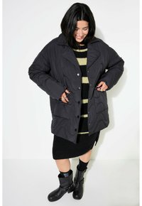 Quiltet sort pufferjakke med en stor krave, trykknapper og sidelommer, båret over en stribet sort og beige sweater og sort nederdel.