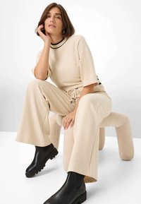 Top beige a maglia con scollatura nera, pantaloni beige a gamba larga abbinati e stivaletti neri con suola texturizzata. Modella seduta su uno sgabello curvo.