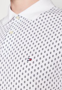 Vit polotröja med en strukturerad tyg och ett upprepat mönster av marinblå prickar, två knappar och en liten broderad logotyp i röd, vit och blå.