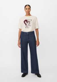 Camiseta gráfica beige con texto y diseño de corazón, combinada con jeans de mezclilla oscuros de pierna ancha y cintura alta. Zapatos negros completan el atuendo.