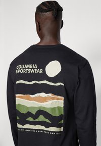 Férfi fekete hosszú ujjú pólóban, hegyvidéki tájjal, nappal és a hátán a "Columbia Sportswear" felirattal.