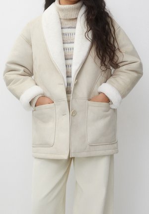 Femme portant un manteau en peau lainée écru avec de grands revers, des poches avant et des poignets retroussés, par-dessus un pull à col roulé beige à motif.