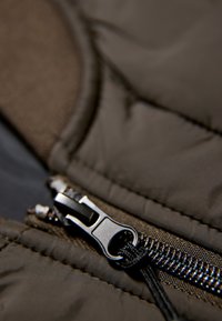 Mörkbrun puffjacka i tyg med en främre dragkedja som har en svart draggrepp och texturerade detaljer som framhäver sömsydda mönster och sömmar.