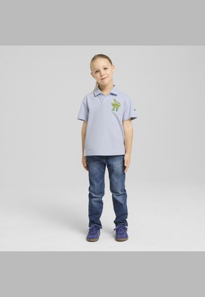 Ljust blå pikétröja med korta ärmar och en grönmönstrad dinosauriebroderi, tillsammans med blå jeans och lila sneakers.