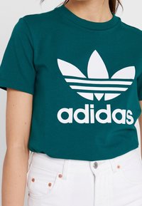 Turkusowa bawełniana koszulka z ribbed crew neck, z dużym białym logo Adidas i trzema poziomymi paskami na całym wzorze.