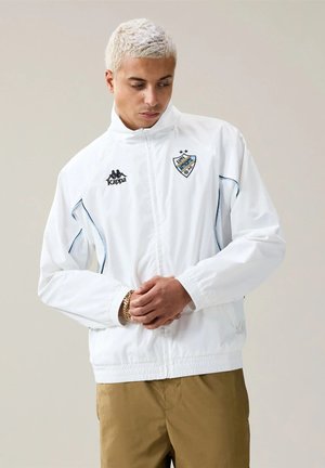 Persona con capelli biondi corti che indossa una giacca bianca Kappa con zip e loghi e pantaloni beige, in piedi davanti a uno sfondo neutro.