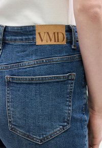 Κοντινή λήψη της πίσω τσέπης από μπλε τζιν με καφέ δερμάτινη ετικέτα μάρκας VMD και μέρος του χεριού ενός ατόμου και λευκό τοπ ορατά.