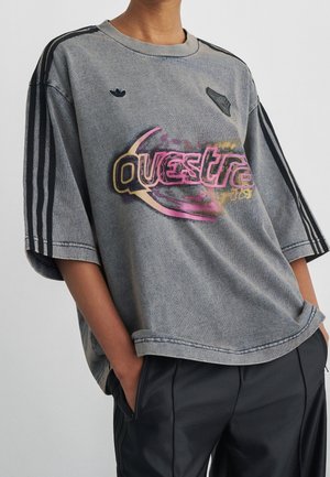 Person iført en falmet grå oversized T-shirt med sorte striber på ærmerne og farverig grafisk tekst "Questra" foran.