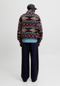 Chaqueta de forro polar con patrones geométricos en rojo, azul marino y crema; combinada con una camisa de mezclilla y pantalones de pierna ancha oscuros. Se muestra la vista trasera.