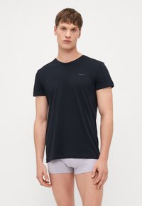 Joven con una camiseta negra ajustada de manga corta y calzoncillos tipo bóxer gris claro, de pie frente a un fondo blanco liso.