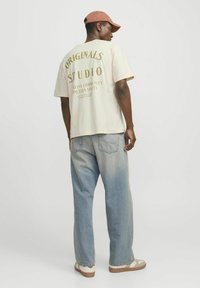 Crème t-shirt met korte mouwen en gouden tekst op de achterkant, gecombineerd met vervaagde blauwe jeans en een lichtbruine pet. Gestructureerde stof en een ontspannen pasvorm.