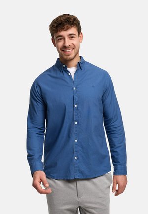 Hymyilevä nuori mies kiharat hiukset, sininen napitettava paita valkoisen paidan päällä ja harmaat housut, seisoo yksinkertaisen taustan edessä.