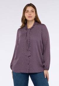 Femme aux cheveux châtain clair portant un chemisier à manches longues avec un motif et un nœud au col, ainsi que des jeans bleus, se tenant contre un fond blanc.