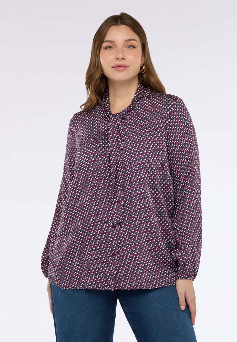 Femme aux cheveux châtain clair portant un chemisier à manches longues avec un motif et un nœud au col, ainsi que des jeans bleus, se tenant contre un fond blanc.