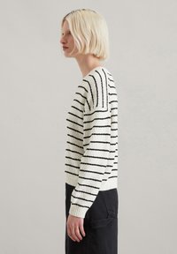 Weißer Pullover mit schwarzen horizontalen Streifen, aus strukturiertem Strick, mit gerippten Bündchen und entspannter Passform. Kombiniert mit schwarzen Hosen.