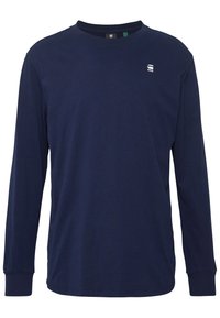 T-shirt à manches longues bleu marine en coton, avec un col rond, des poignets ajustés et un petit logo blanc sur la poitrine. Texture douce.