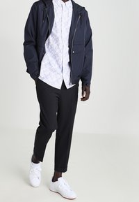 Navy windbreaker with a drawstring hood, white floral-patterned shirt, black tapered pants, and white sneakers.