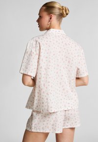 Vrouw met een blonde bun, gekleed in een wit shirt met korte mouwen en bijpassende korte broek met een klein roze bloemmotief, gezicht naar weg van de camera.