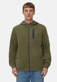 Haut à capuche zippé vert olive, avec une capuche, une poche zippée noire sur la poitrine et des poches latérales. Fabriqué dans un tissu doux et texturé.