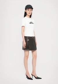 Biela tričko s čiernym logom, skombinované s hnedou koženou mini sukňou s zipsami. Čierne vysoké topánky dopĺňajú outfit.