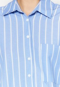 Ljusblå skjorta med knappar och vita vertikala ränder, med krage, bröstficka och vita knappar. Slät, vävd textur.