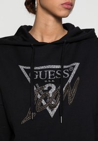 Black hooded sweatshirt with a triangular graphic featuring the text "GUESS" and "ICON" in cracked silver and textured metallic accents.