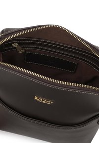 Hnedá kožená taška so zlatými kovovými súčiastkami, ktorú predný zips otvára do hlavného oddelenia a obsahuje aj vnútornú kapsu. Okolo okrajov sú vyšité detaily.