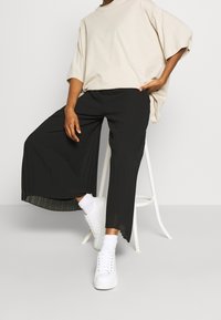 Zwarte plooibroek gecombineerd met een losse beige top, aangevuld met witte sneakers en enkelsokken. Kenmerkend is de ontspannen pasvorm en zachte texturen.