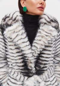 Cappotto in pelliccia sintetica con righe bianche e nere, caratterizzato da un ampio colletto e una consistenza morbida. Modella che indossa un dolcevita nero e orecchini circolari verdi.