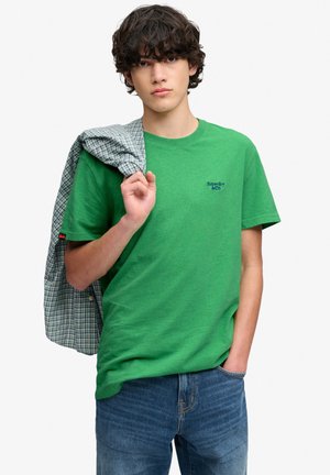 Ung mand med krøllet hår iført en grøn Superdry t-shirt og blå jeans, der holder en blå ternet skjorte over skulderen.