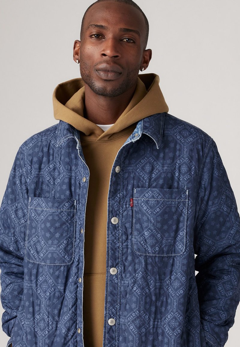 Chaqueta de quilted en color marino con un patrón geométrico, dos bolsillos en el pecho y un cuello de mezclilla en contraste sobre una sudadera color beige. Cierre de botón visible.